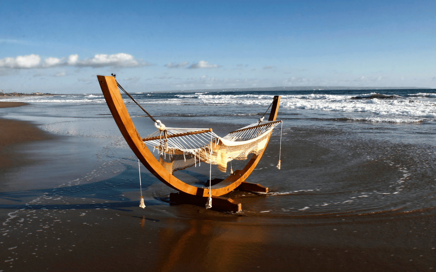 A curved wooden hammock frame with a cream hammock standing in the shallow surf of a beach.