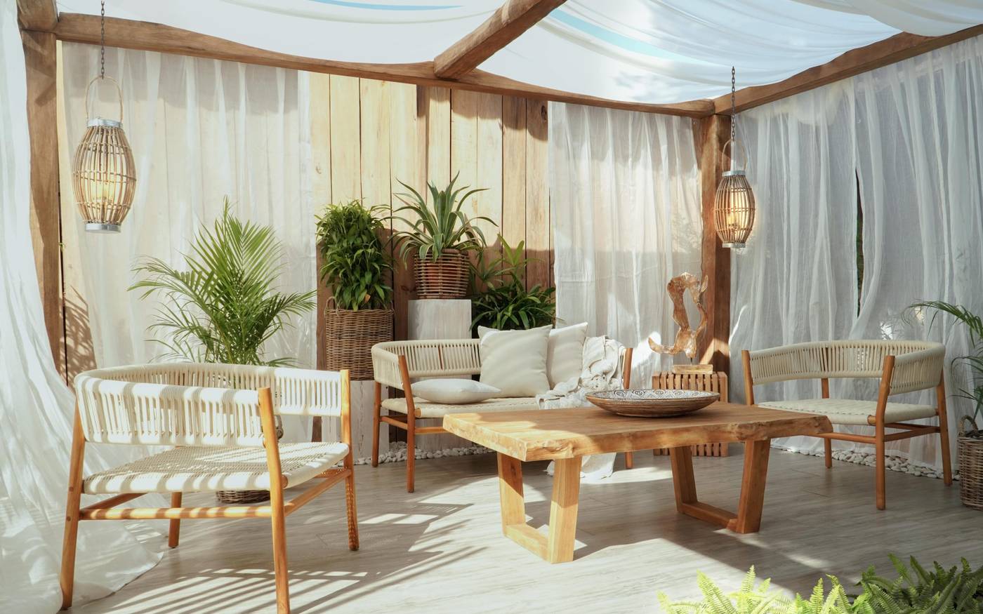 A covered teak terrace with white drapes, two rope-backed benches, a live-edge coffee table and hanging lanterns among palms and ferns.