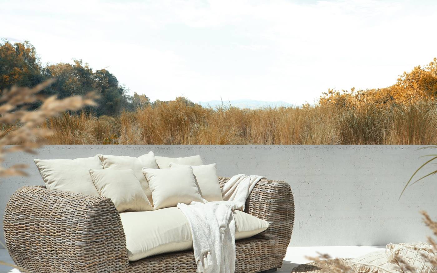 A grey wicker sofa with cream cushions and a white throw on a terrace edge, with a round white rug, dry grass and a tree line behind.