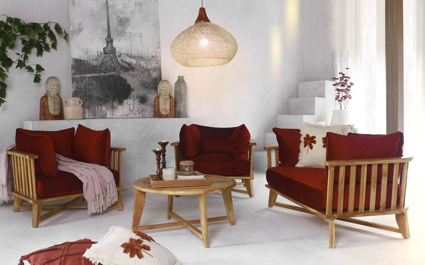 A living room with three teak slatted armchairs with maroon cushions, a round teak coffee table, a Paris painting, two bust statues and a rattan pendant lamp.