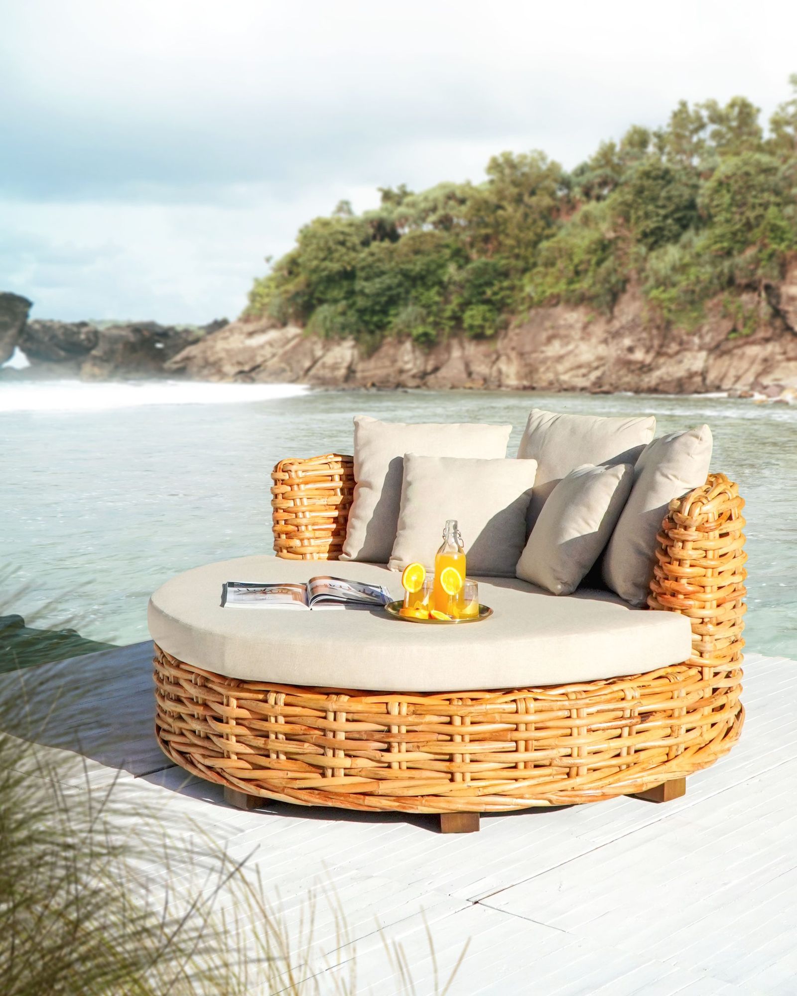 A round rattan daybed with a cream cushion and grey pillows on a white deck above a rocky bay.