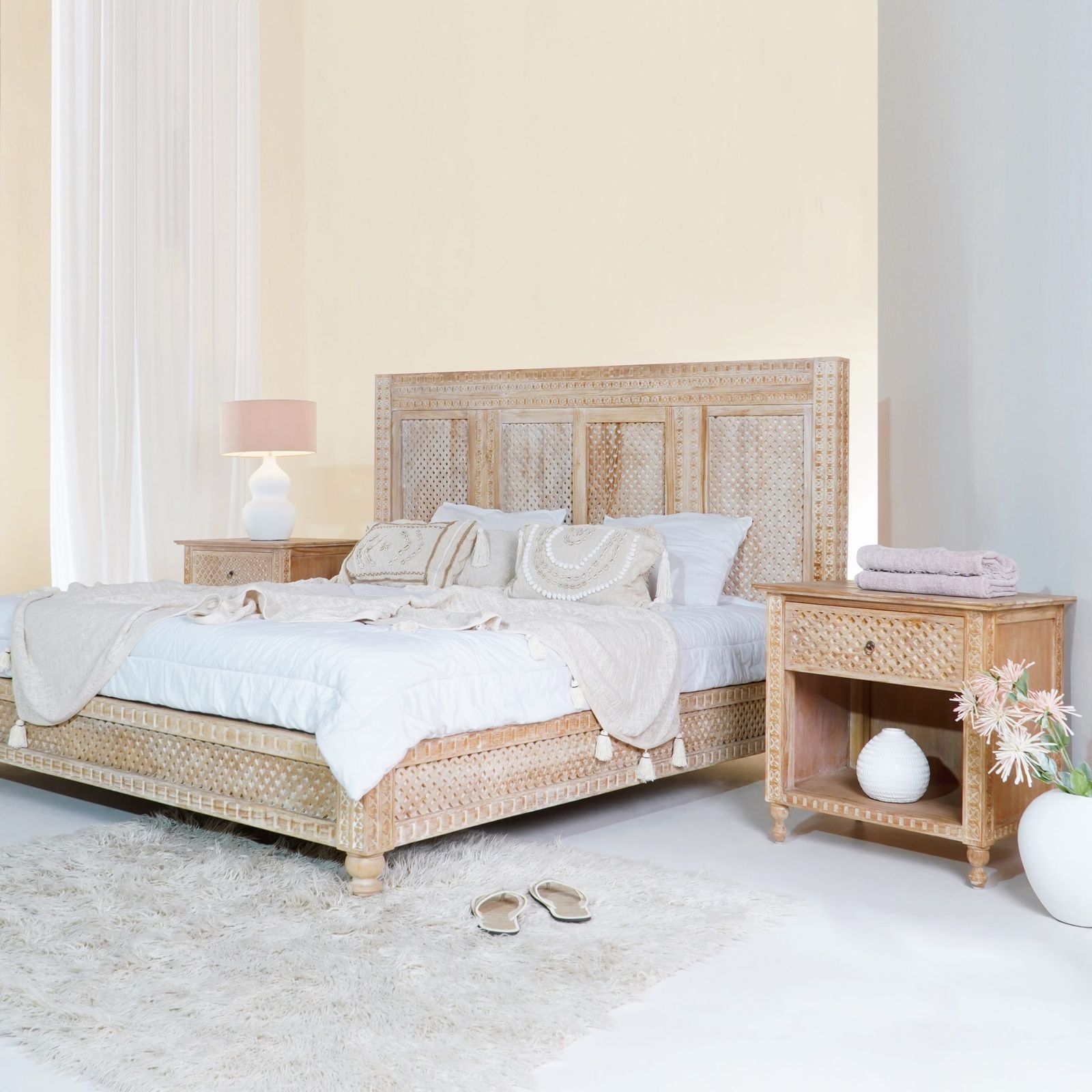 A carved teak bed with a lattice headboard, a matching bedside table, white bedding and a shaggy rug in a bright bedroom.