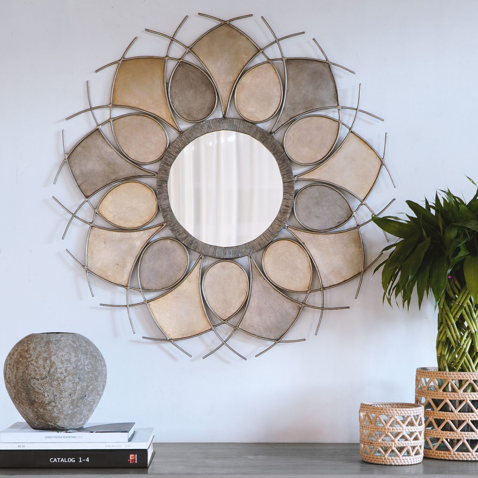 A round flower-shaped metal wall mirror in gold and bronze tones above a shelf with a stone pot, a plant and woven holders.