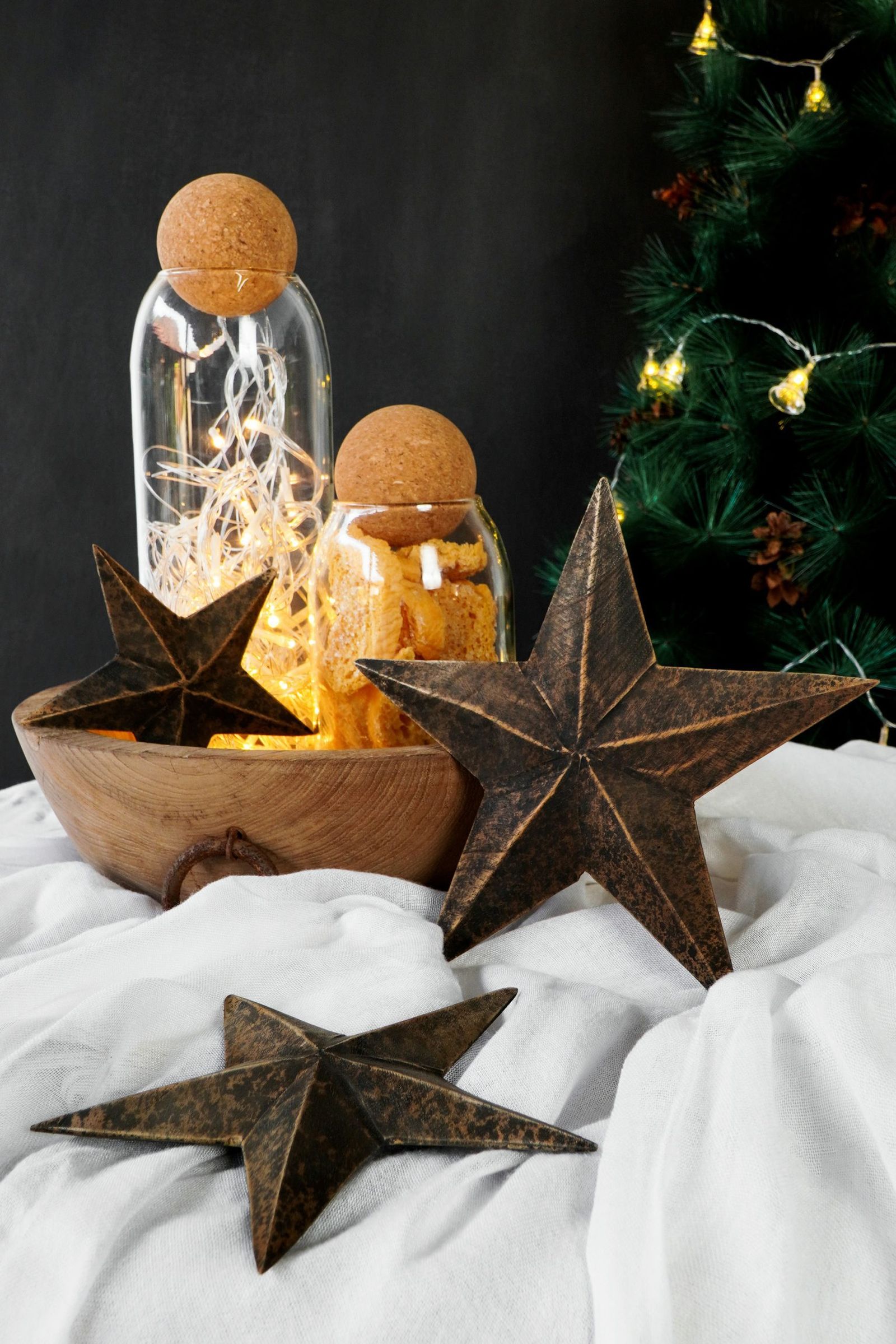 Three rusty-brown metal stars in a wooden bowl on white linen, with glass jars lit by fairy lights and a Christmas tree behind.