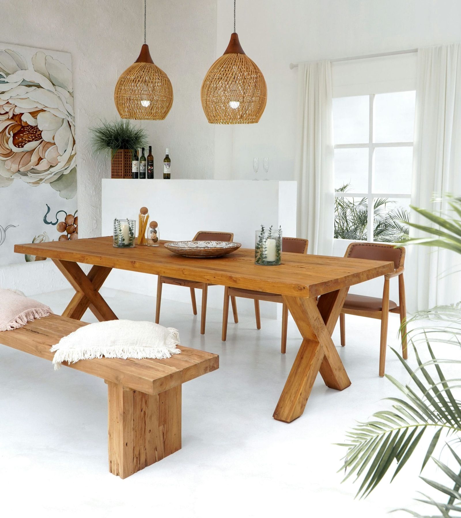 A teak dining table with crossed legs, a thick teak bench and leather dining chairs under two rattan pendant lamps in a white room.
