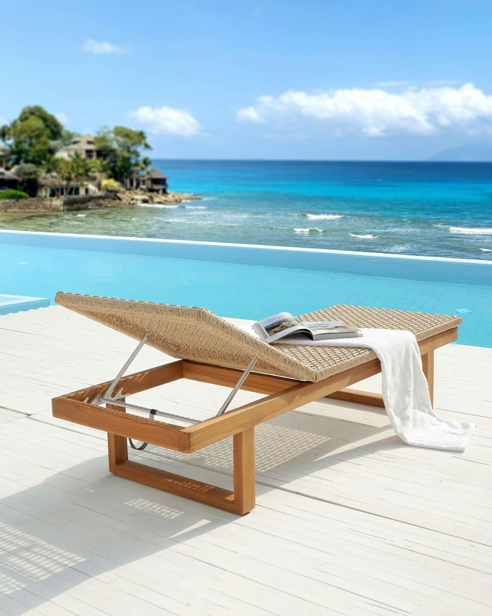 A teak sun lounger with a woven seat and a raised backrest on a white pool deck, with an infinity pool and the sea behind.