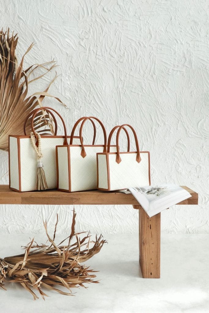 Three white woven handbags with tan leather handles on a thick teak bench, against a plastered wall with dried palm leaves.
