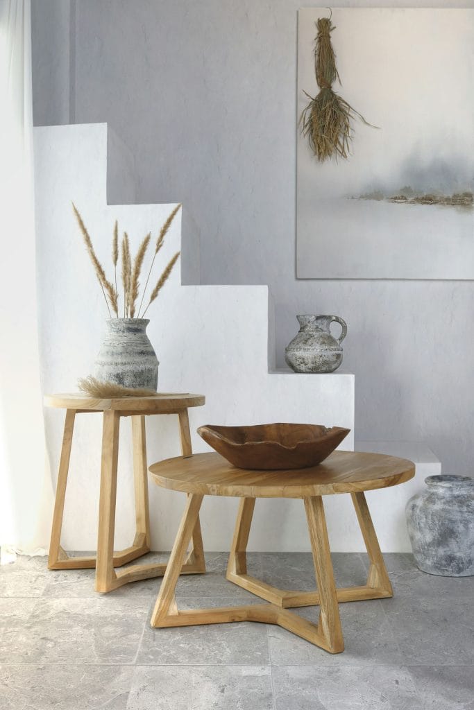 Two round teak nesting tables with a carved wooden bowl, a stone jug and vases against a white staircase.