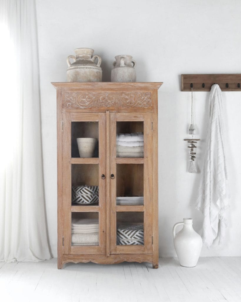 A carved teak glass-door bookcase in a white room, with grey jars on top, towels inside and a white jug beside it.