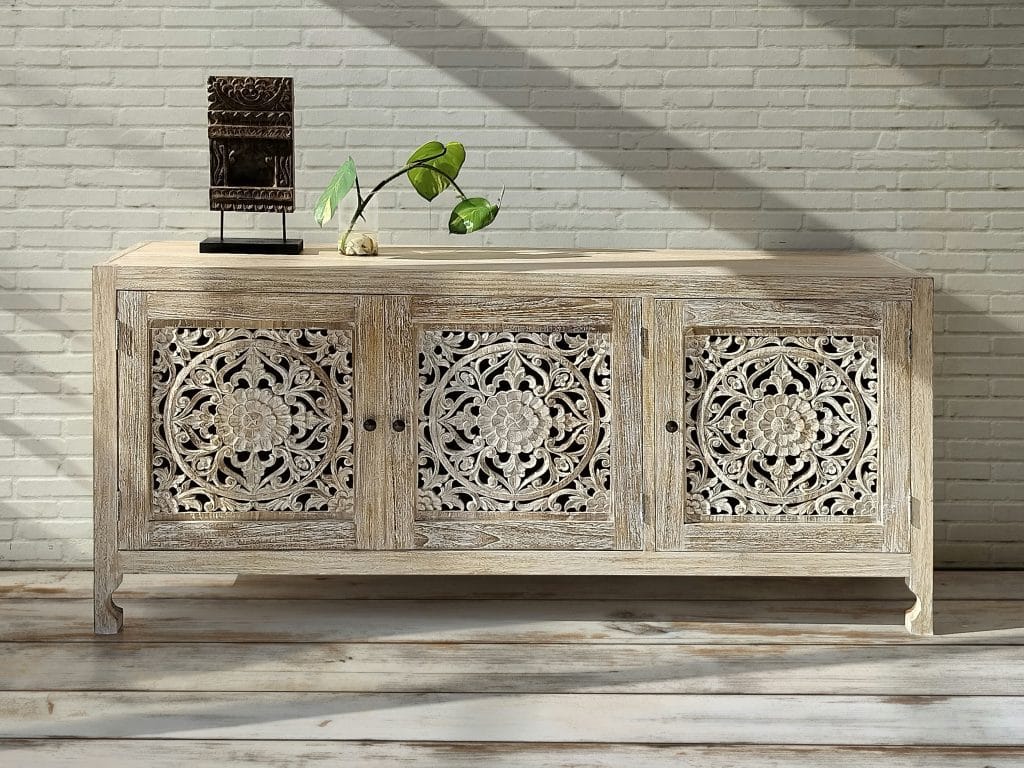A whitewashed teak buffet with three carved mandala doors against a white brick wall, with a small carved ornament and a plant on top.