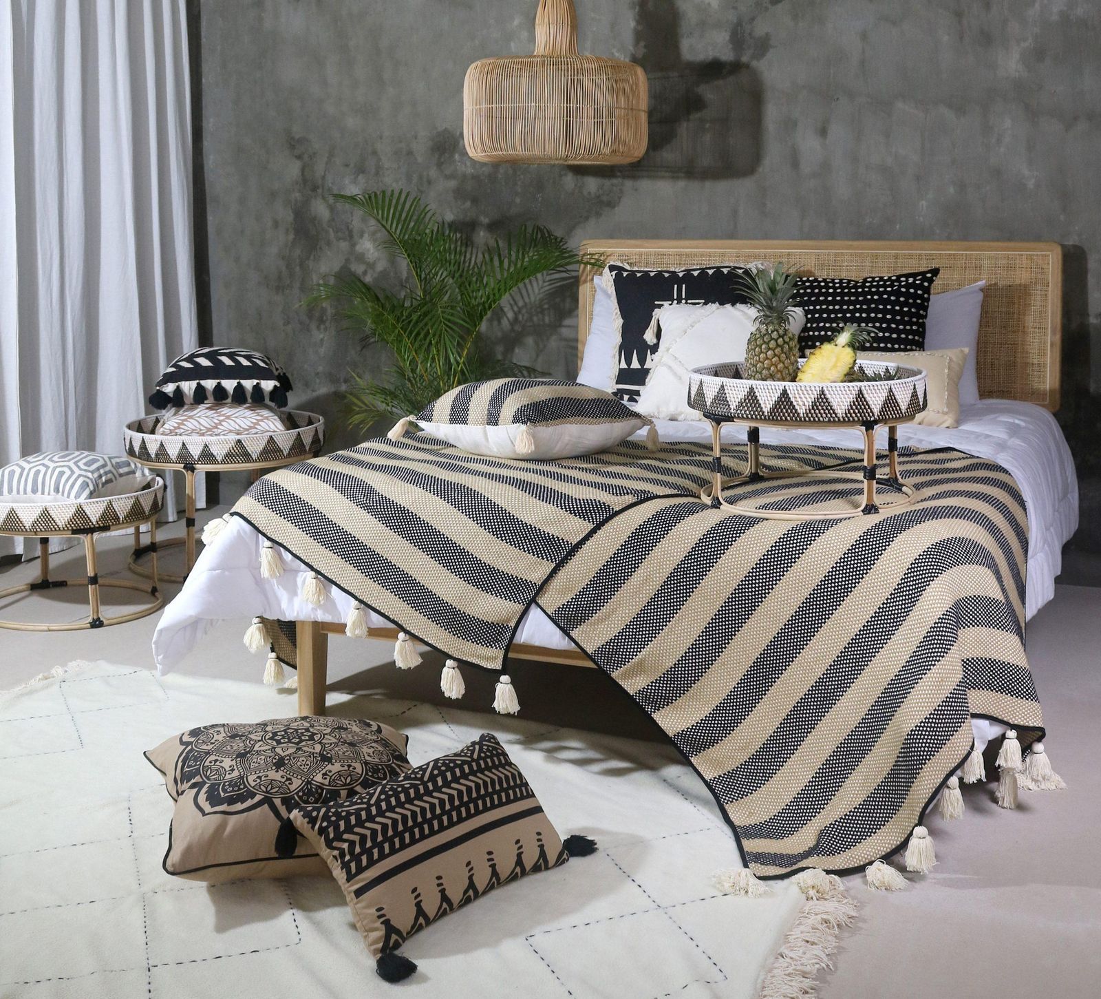 A bed with a rattan headboard, a black and cream zig-zag striped blanket and patterned cushions, with tray tables on a pale rug under a rattan lamp.