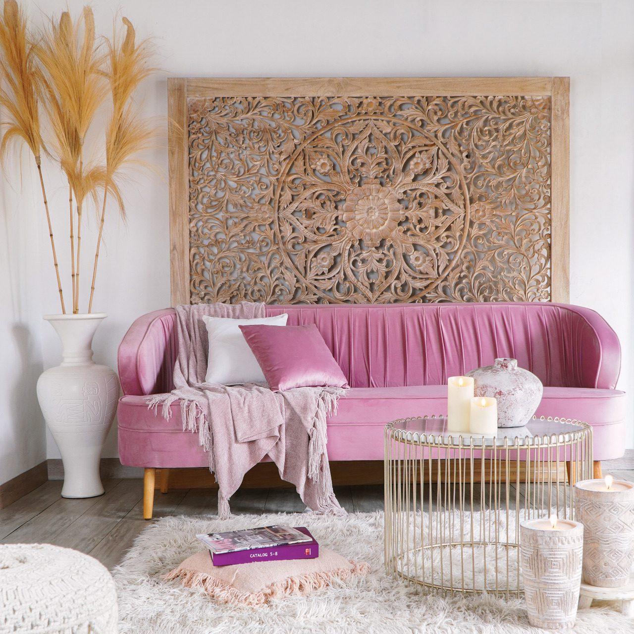 A pink velvet sofa under a large carved wooden panel, with a gold wire coffee table, a tall white vase, dried grasses and candles.