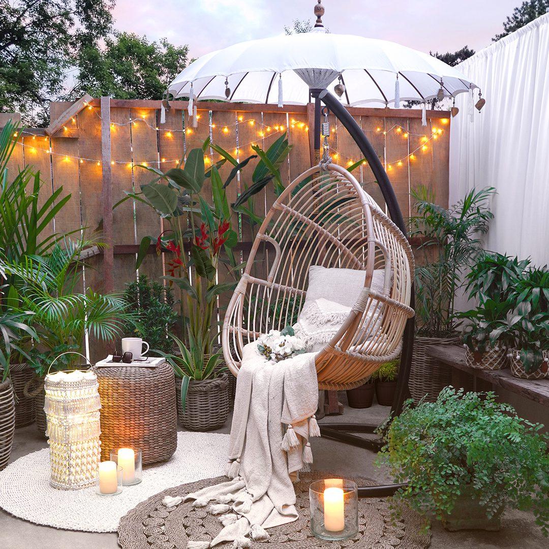 A rattan hanging egg chair with a cream throw under a white Balinese umbrella in a lit garden corner, with candles and lanterns.