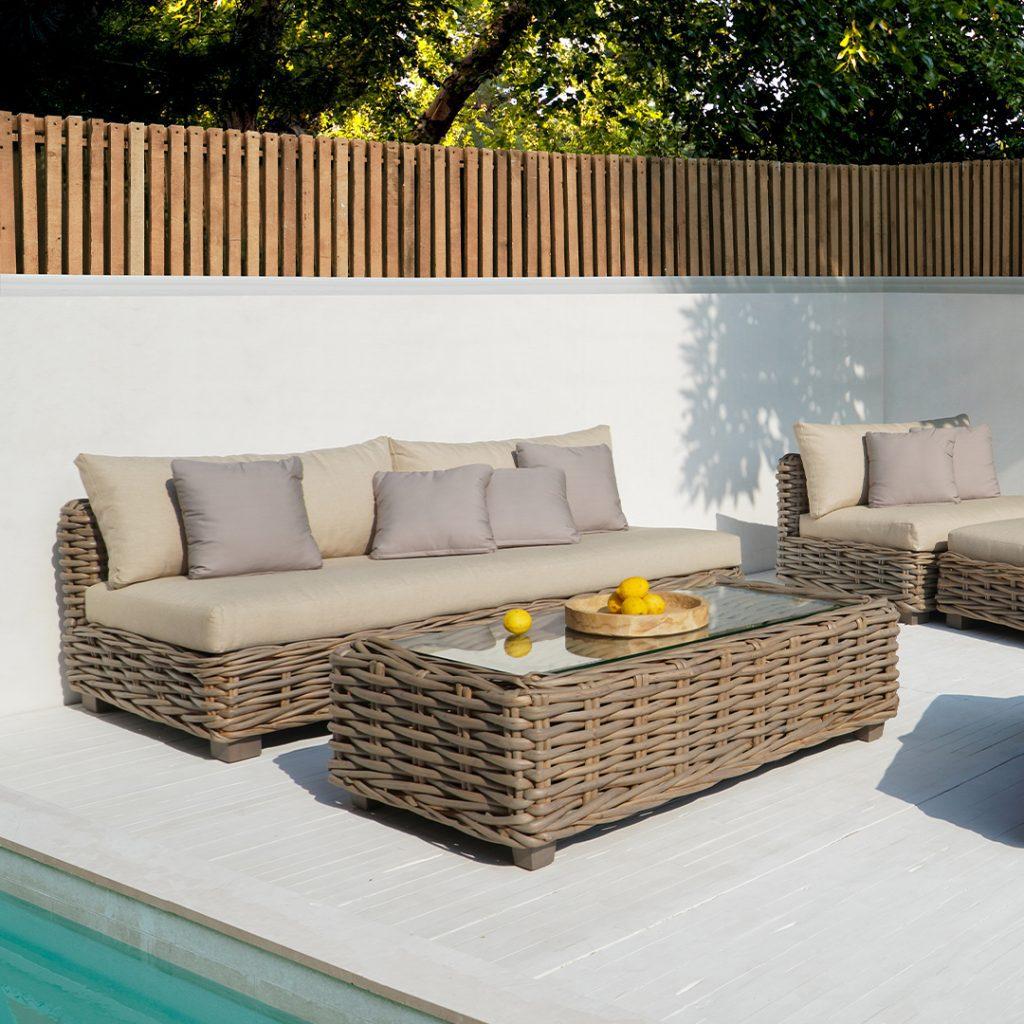 A rattan outdoor lounge set with beige cushions, a long sofa, a corner chair and a glass-top coffee table beside a pool.