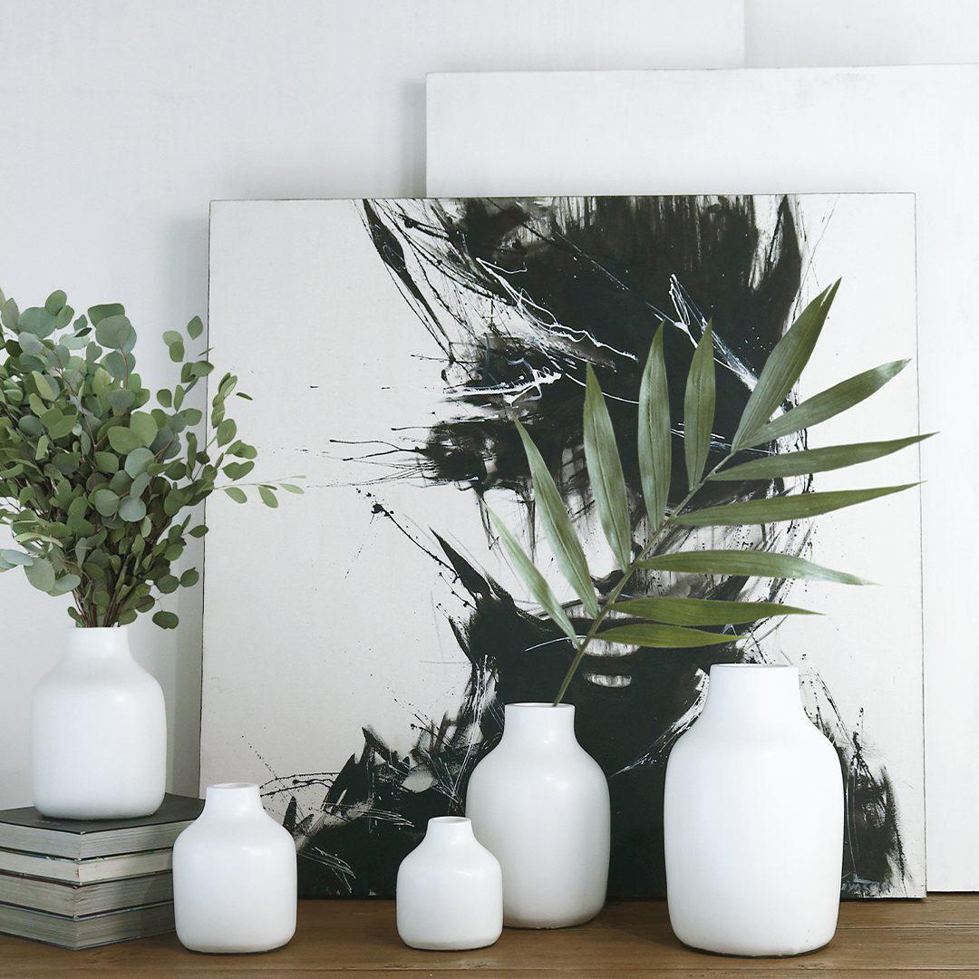 Five white ceramic vases of different sizes in front of a black and white abstract painting, with eucalyptus in the smallest.