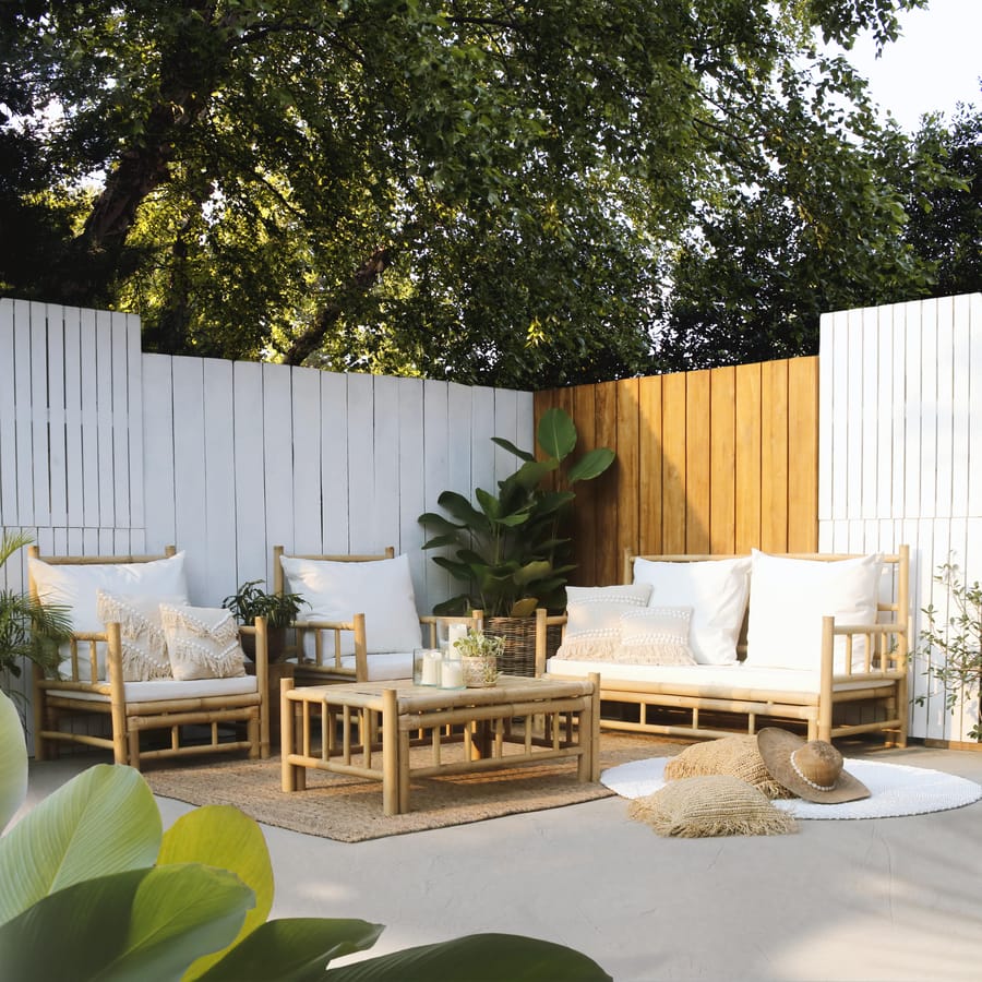 A bamboo outdoor lounge set with a sofa, two armchairs and a low open-sided table with white cushions, in a garden corner with white and wooden fences.